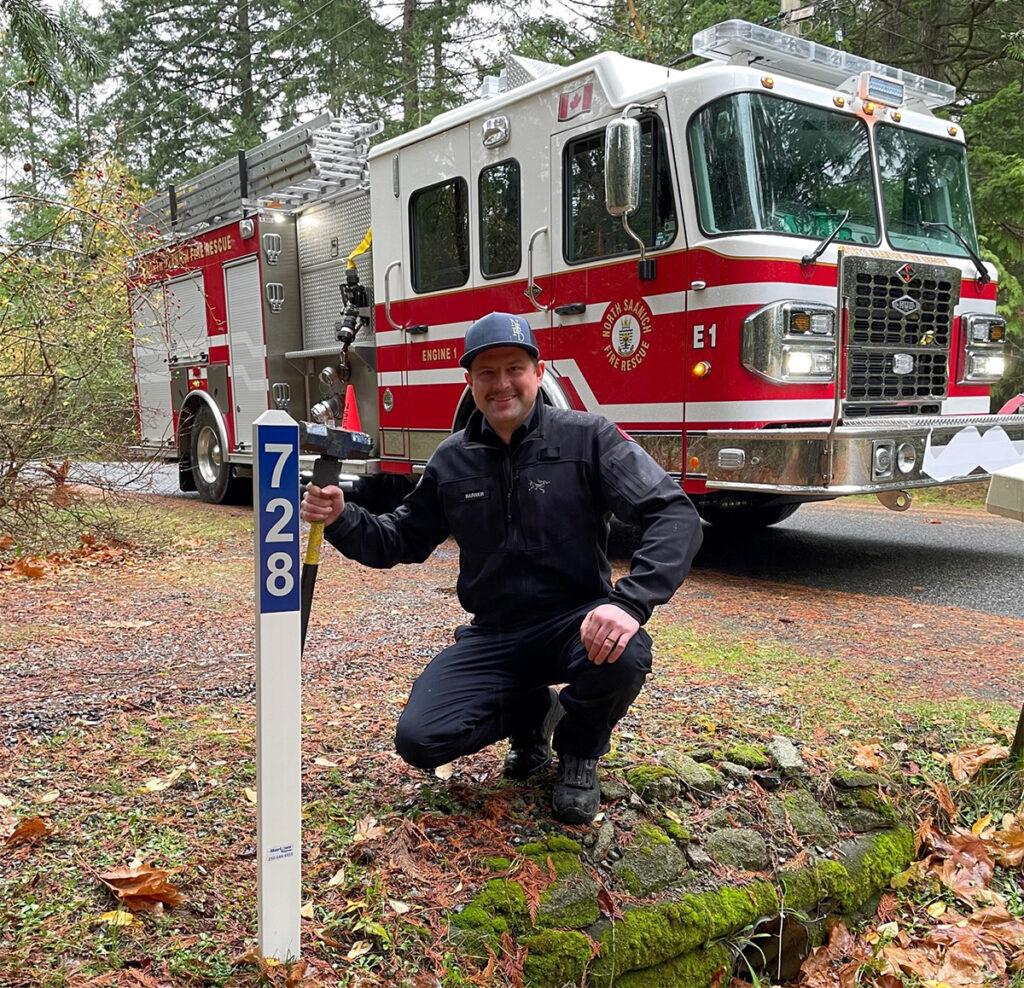 Firefighter crouching beside a blue and white address sign, with a fire truck in the background.