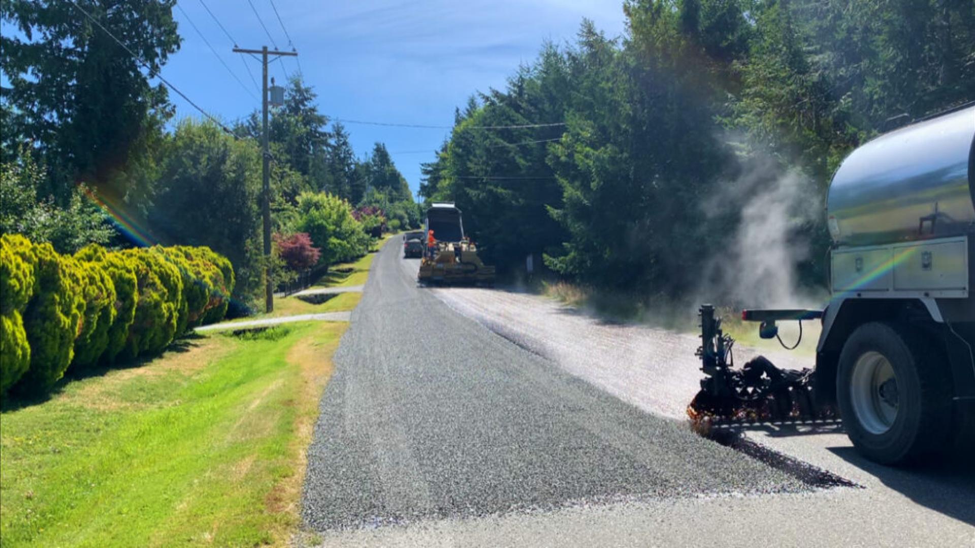 Construction equipment laying gravel on a rural road, surrounded by trees and blue skies.