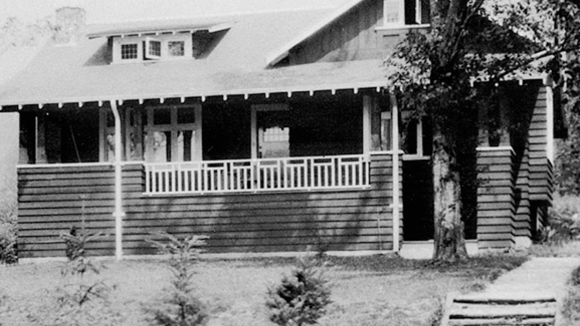 Historic wooden house with a large porch and stone pathway, surrounded by trees.