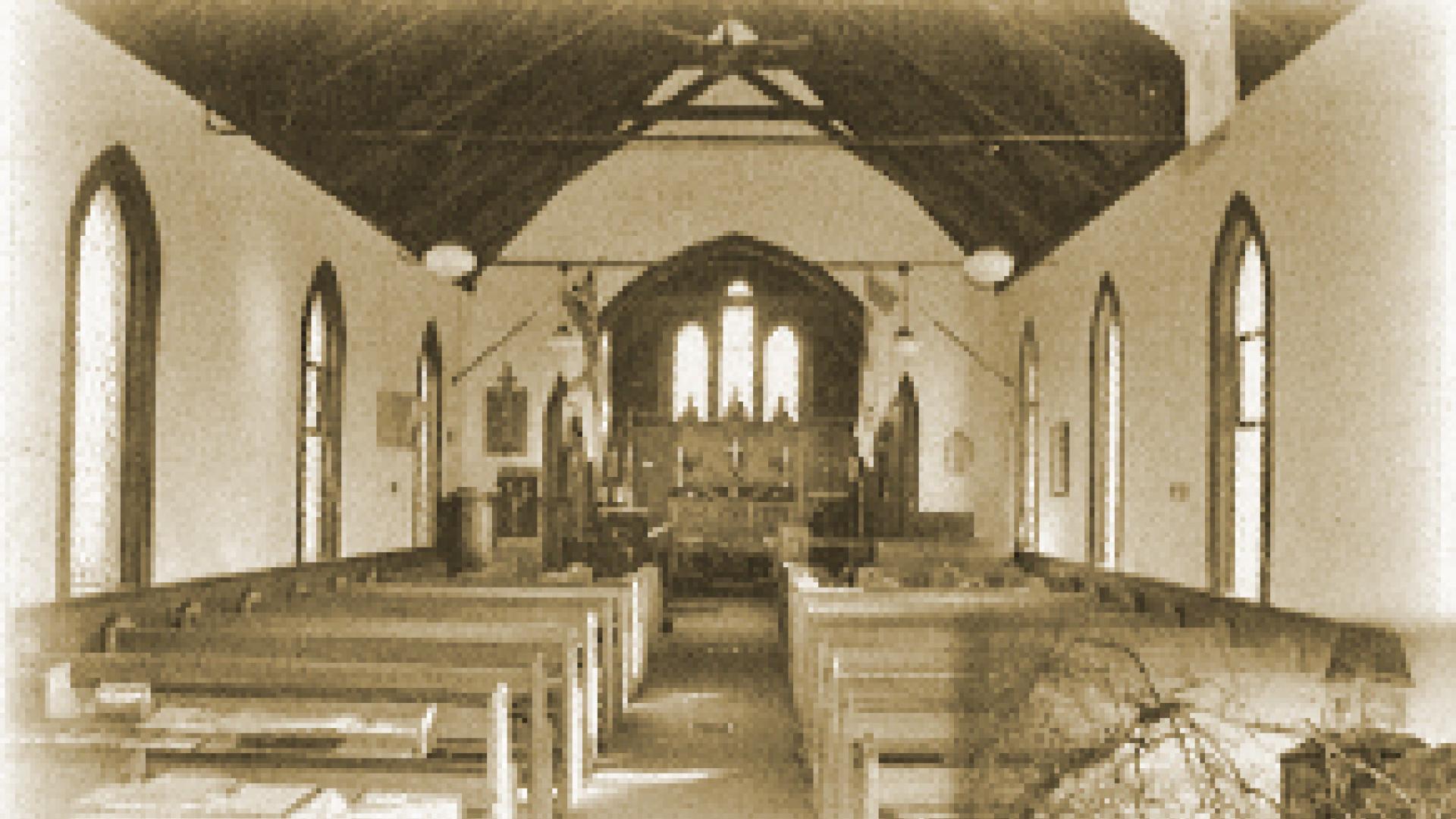 Interior of a rustic church with wooden benches and arched windows, tinted sepia.