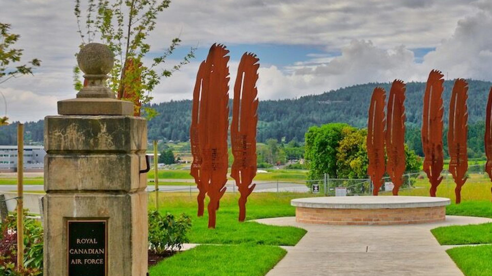 Pathway leading to a circular plaza with metal sculptures and greenery, surrounded by hills.