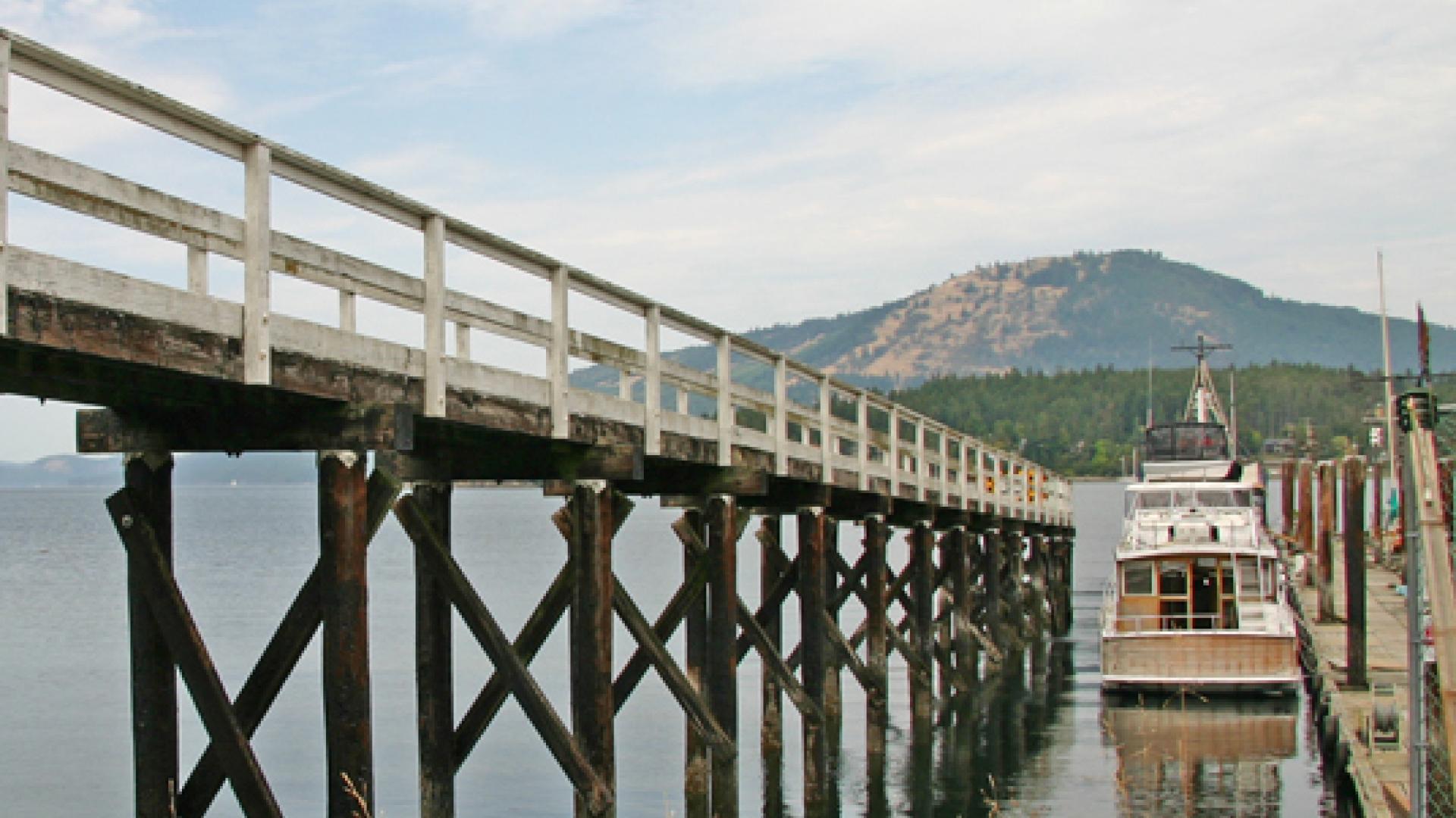 Wooden pier extending over calm water, with a boat docked nearby and mountains in the background.