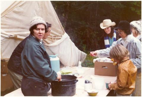 In front of their camping tent are several folks around a table beginning to make dinner.