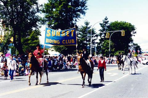 A parade of people walking down a main street. Two people on horseback are in the foreground, holding up a sign that says Sunset Riding Club.