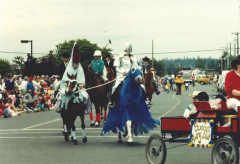 Horses and riders dressed in medieval garb walking along the road for the parade. Lots of spectators sitting at the side of the road watching.