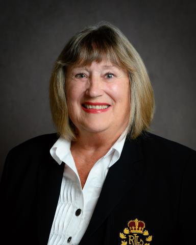 A smiling individual in a dark blazer with a crest, against a grey background.