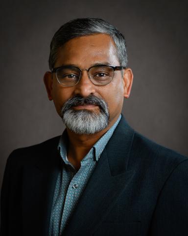 A person with glasses and a beard, wearing a dark blazer, smiles against a dark background.