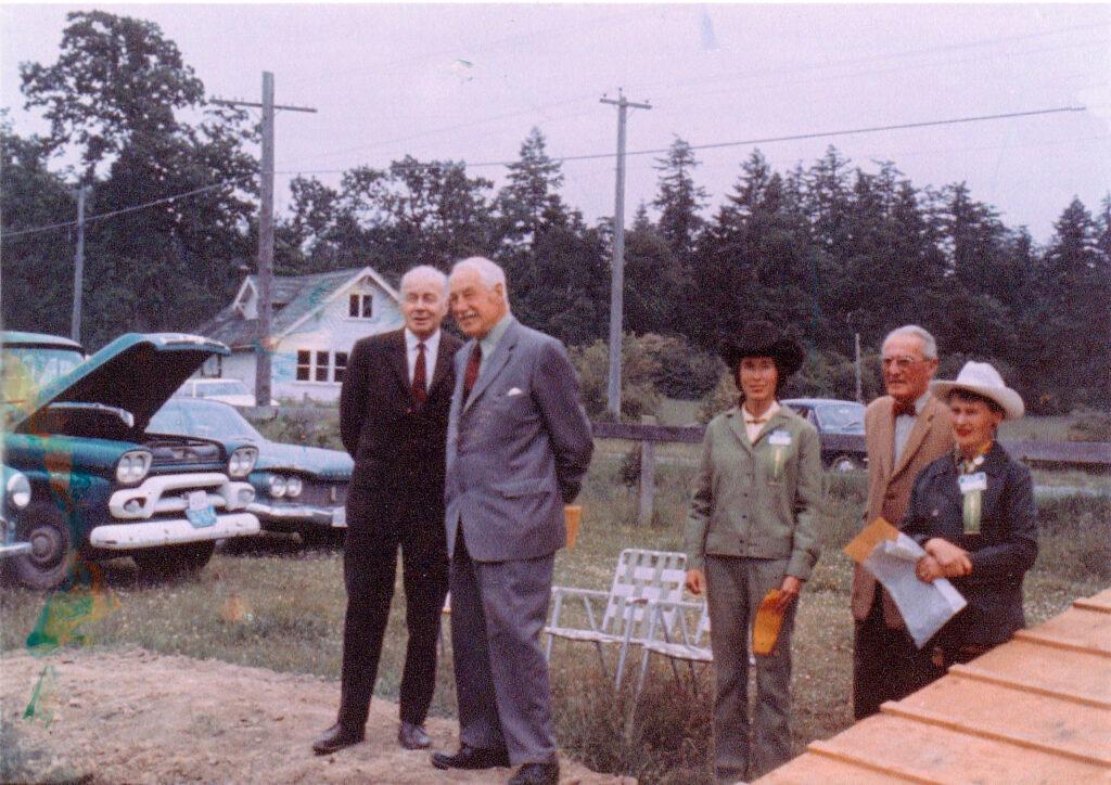 In the foreground two men in suits and ties are smiling. To their left are two women in cowboy hats and an older man in a suit and bowtie. Two vintage cars are visible to the left. The scene is rural residential with open fields, powerlines and trees in the background.