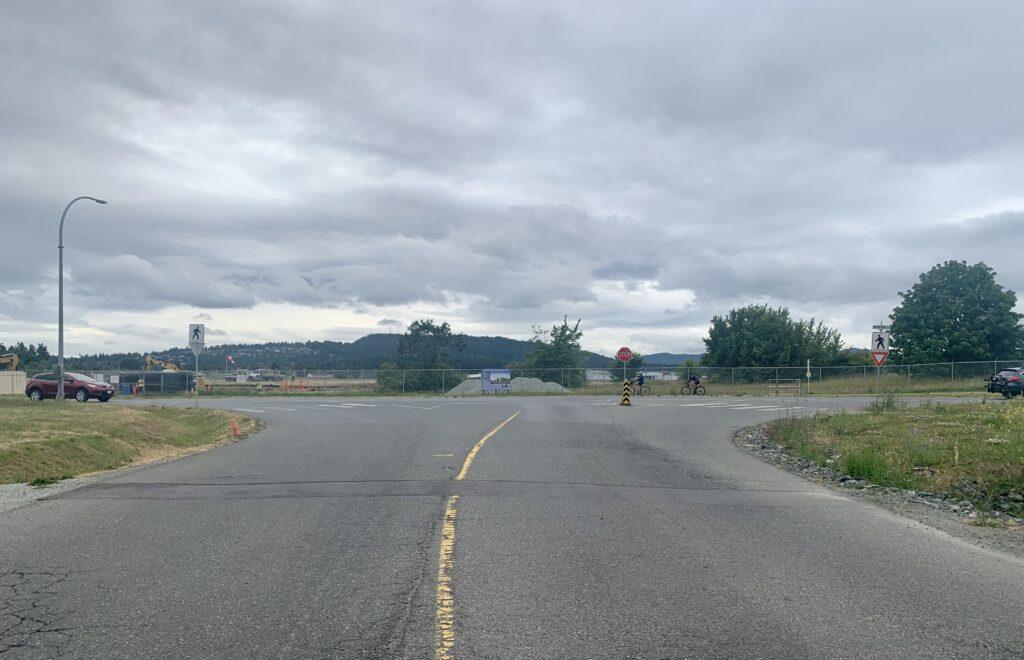 Roadway intersection under a cloudy sky, with a person walking and pastel greenery nearby.