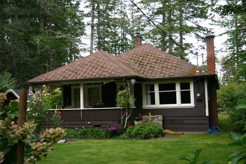 Bungalow surrounded by greenery, featuring a brown exterior and a unique shingle roof.