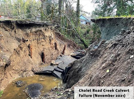 Erosion near a creek with exposed culvert and a damaged road, surrounded by trees.