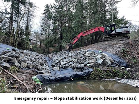 Excavator working on slope stabilization with rocks and a tarp by a stream and trees.
