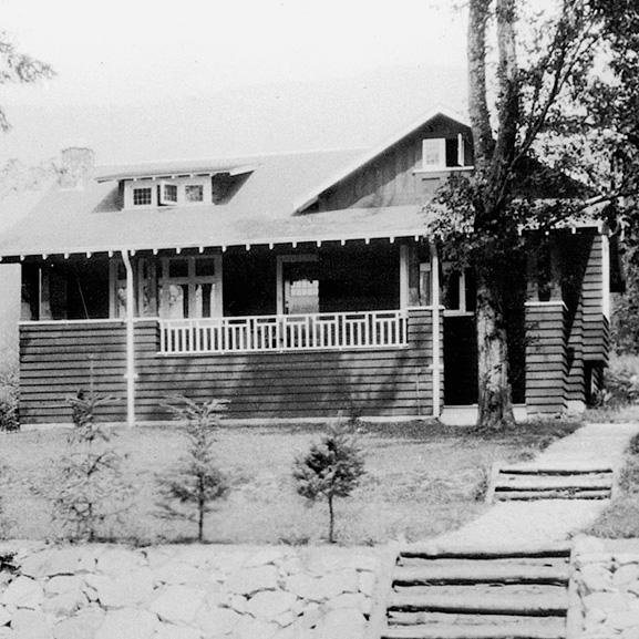 Historic wooden house with a large porch and stone pathway, surrounded by trees.