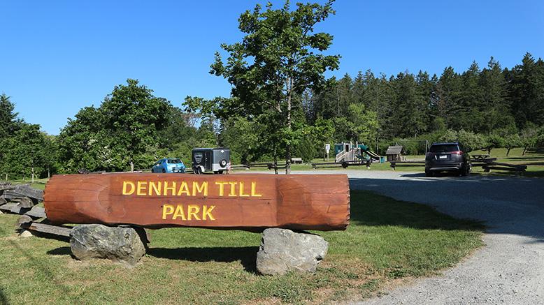 Sign for Denham Till Park on a sunny day with greenery and parked vehicles.