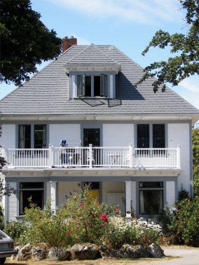 White two-storey house with a grey roof and balcony, surrounded by flowering bushes.