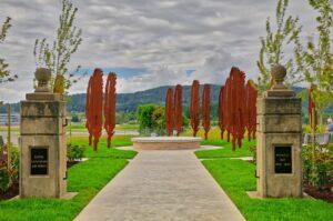 Pathway leading to a sculpture garden, flanked by stone pillars and greenery, under a cloudy sky.