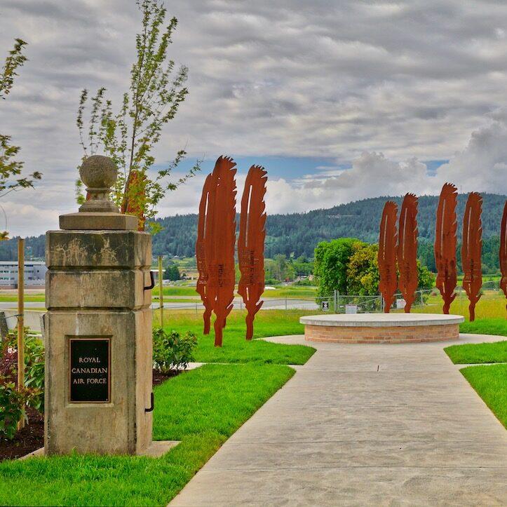 Pathway leading to a circular plaza with metal sculptures and greenery, surrounded by hills.