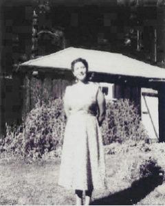 A person stands in front of a small building, surrounded by greenery in a black and white photograph.