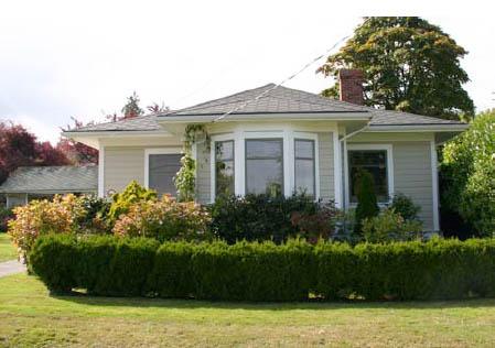 Bungalow with a garden, surrounded by a hedge and trees under a clear sky.