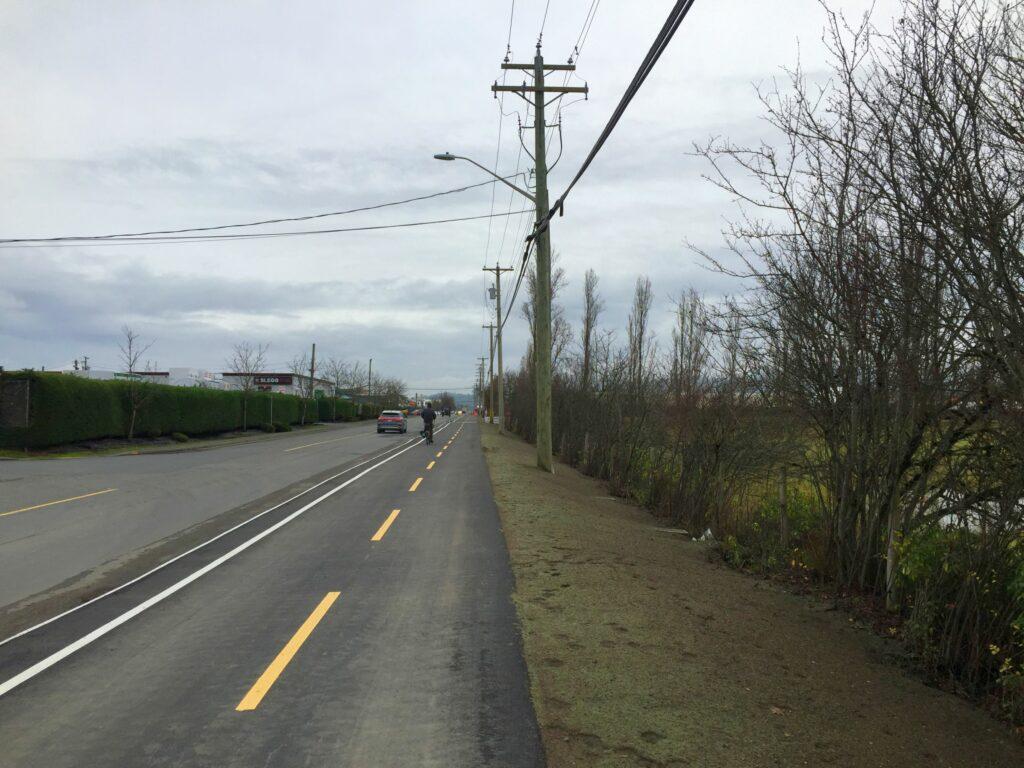 A wide view of a road with power lines, trees, and a cloudy sky.