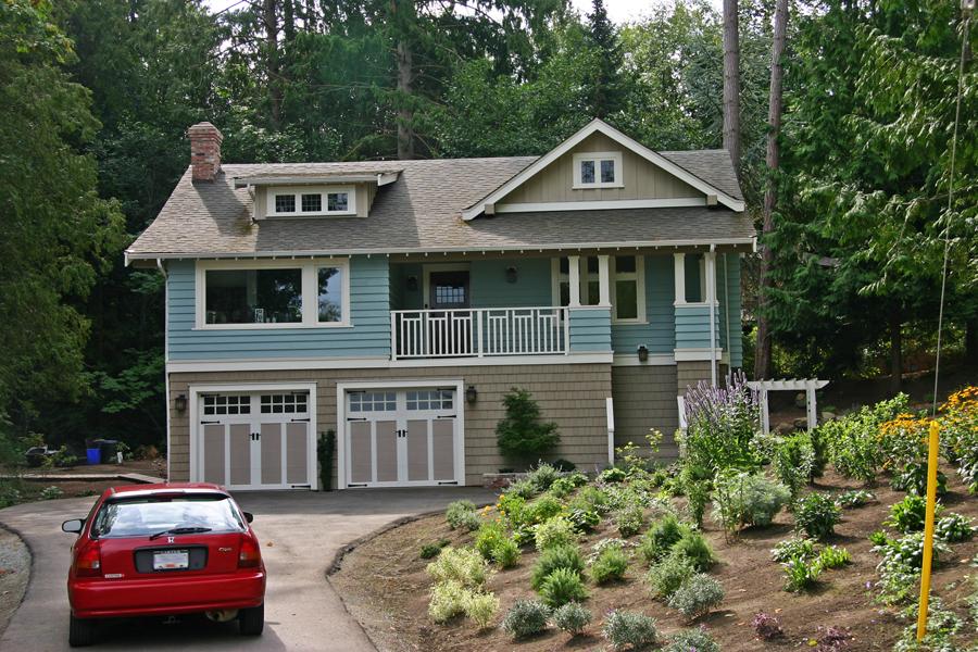 Two-storey blue and beige house with a garage, surrounded by greenery and a red car parked nearby.