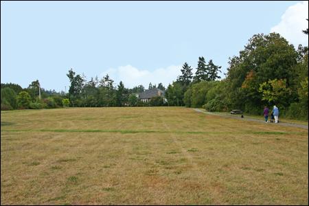 Large open field with a house in the far distance, and the field is surrounded by green trees. It looks like summer because the grass is brown.