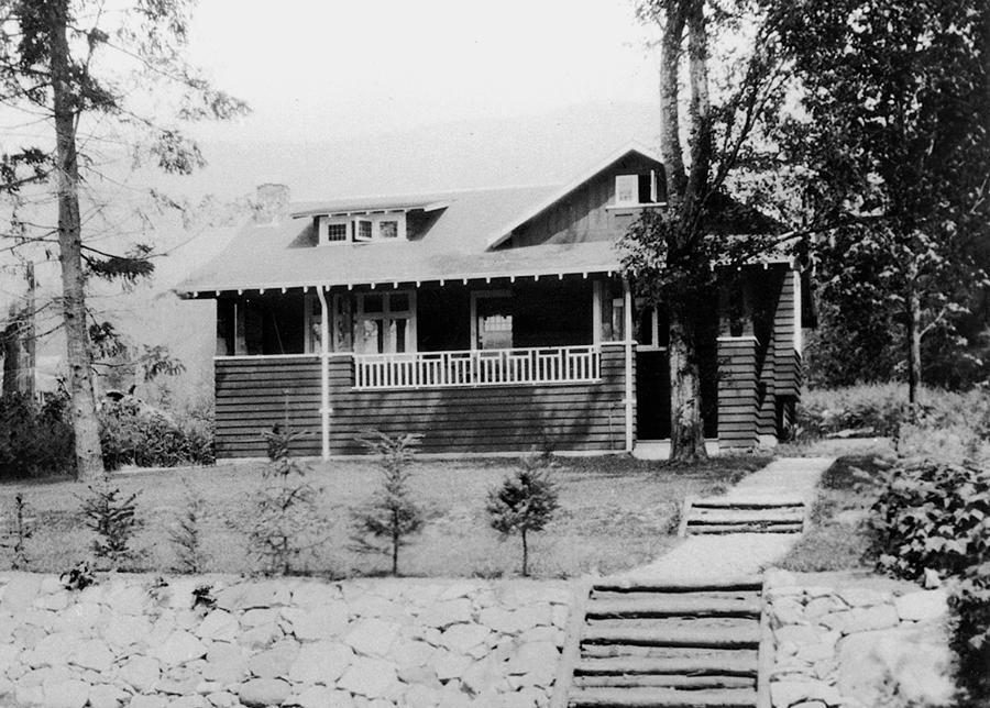 Black and white photograph of a wooden house surrounded by trees and a stone pathway.