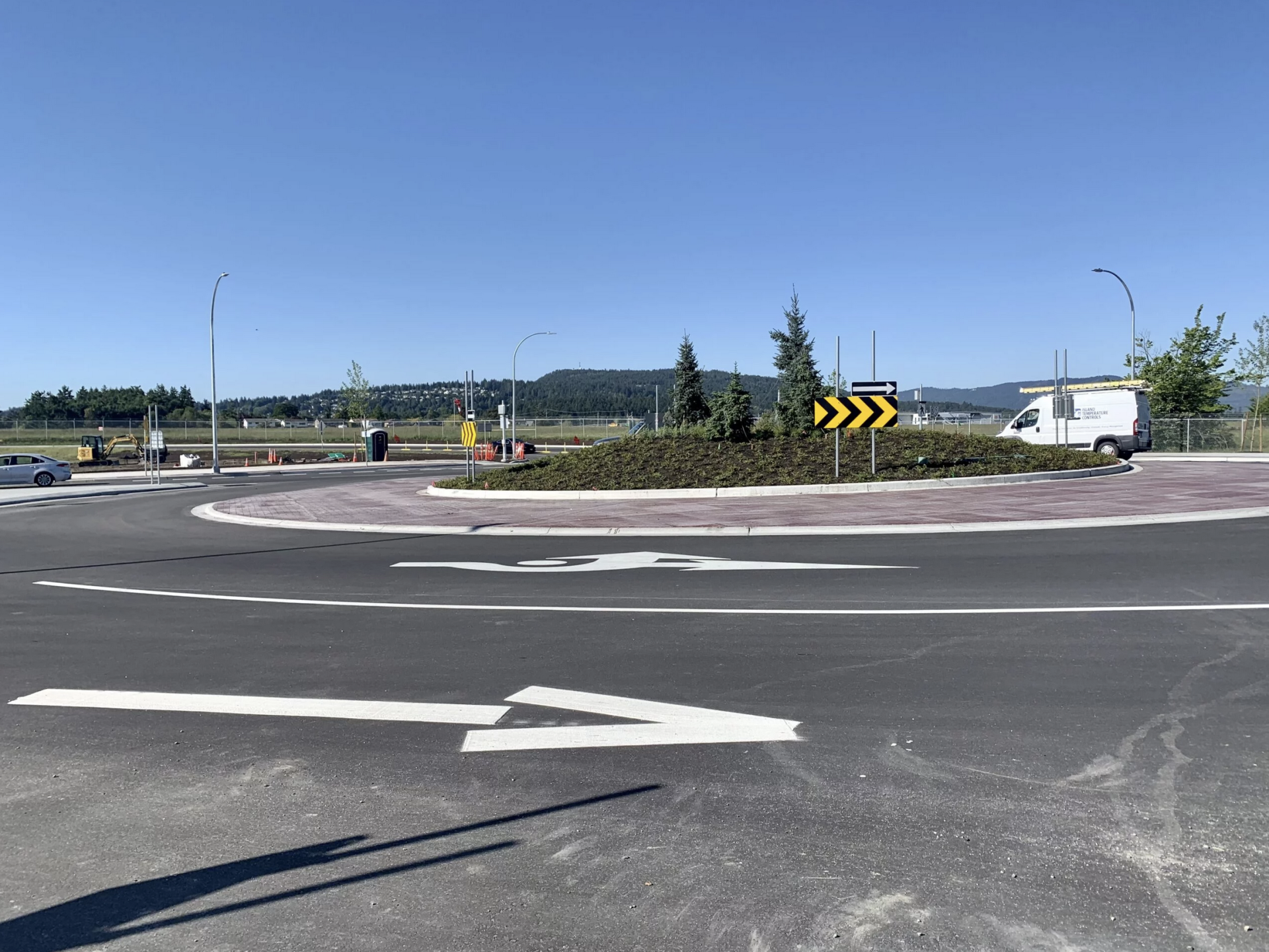 A sunny view of a roundabout with directional signs and parked vehicles in a suburban area.