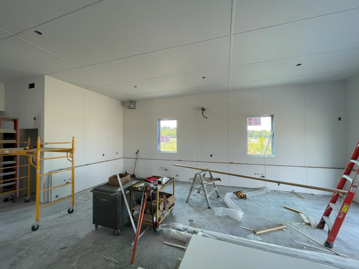 White wall and ceiling panels are installed, while scaffolding, a ladder, work tables, and building materials are positioned throughout the room on the unfinished concrete floor.