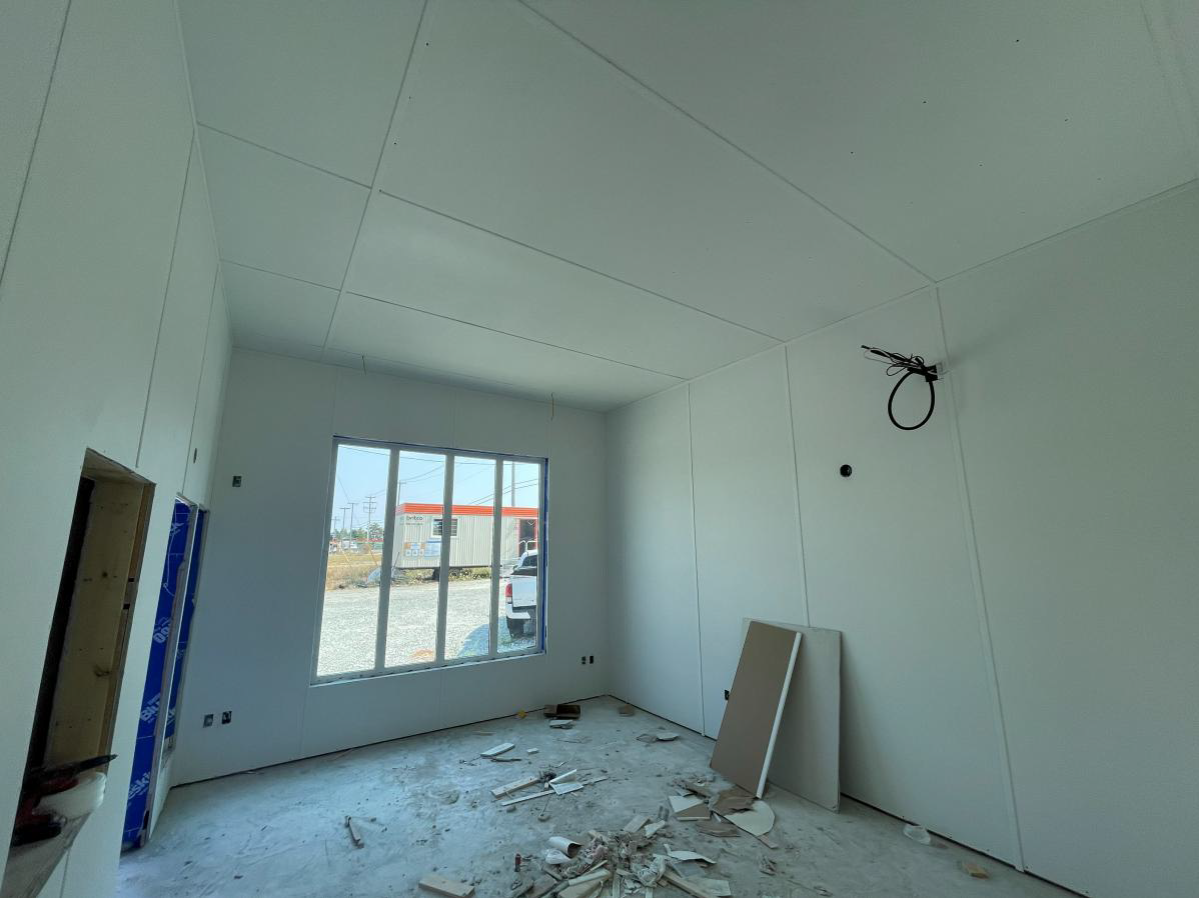 White insulated wall panels and suspended ceiling panels have been installed, with a large front window providing natural light. Construction materials and debris remain on the unfinished concrete floor.